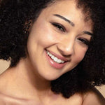A young woman with curly dark hair and clear skin smiles warmly at the camera, wearing vegan makeup from the 100% Pure Fruit Pigmented® Berry Naked Palette 9g by 100% Pure against a neutral background.