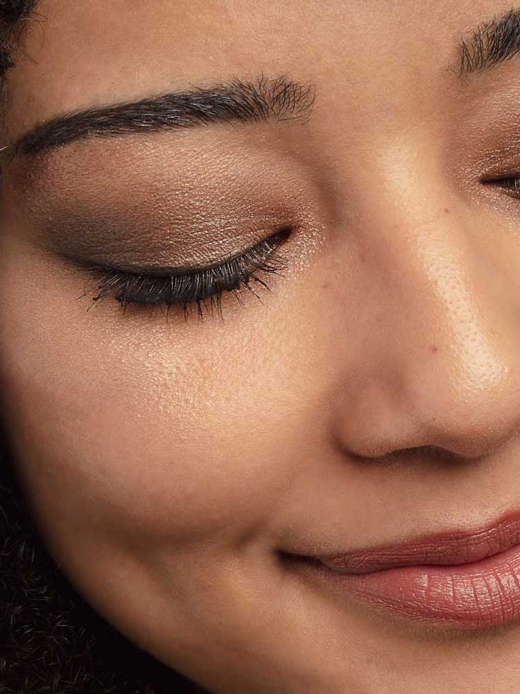A close-up of a person’s face with closed eyes, showcasing shimmery brown tones from the 100% Pure Better Naked Palette 9g, black eyeliner, long lashes, defined brows, and a soft dimpled smile.