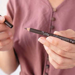 A person in a mauve buttoned shirt holds the 100% Pure Creamy Long Last Liner 1.14g by 100% Pure in one hand and its cap in the other, getting ready to apply the creamy eyeliner.
