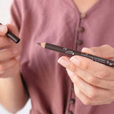 A person in a mauve buttoned shirt holds the 100% Pure Creamy Long Last Eye Liner 1.14g in one hand and its cap in the other, ready to apply or store this long-lasting eyeliner.