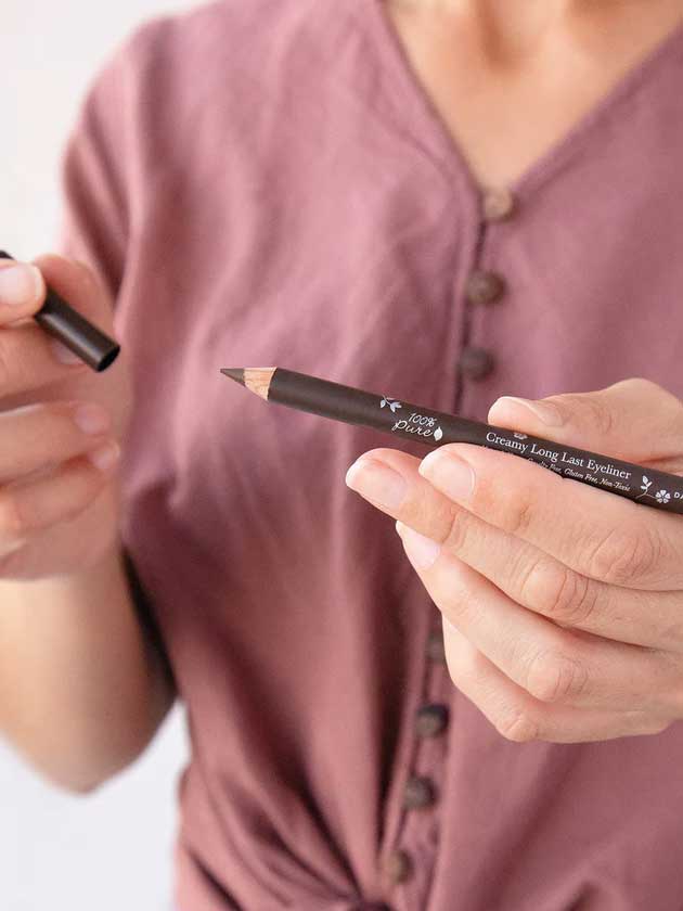 A person in a mauve buttoned shirt holds the 100% Pure Creamy Long Last Eye Liner 1.14g in one hand and its cap in the other, ready to apply or store this long-lasting eyeliner.