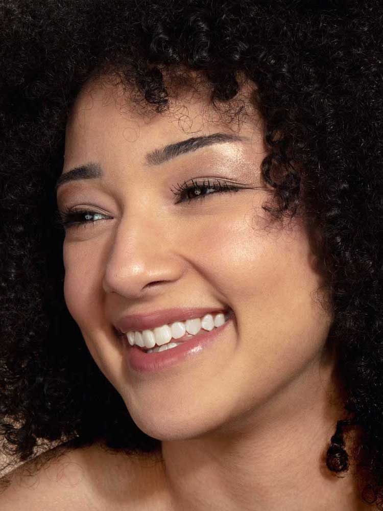 A young woman with curly dark hair and clear skin smiles brightly, wearing natural vegan makeup from the 100% Pure Pretty Naked Palette 9g. She looks slightly to the side, radiating joy.