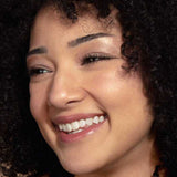 A young woman with curly dark hair and clear skin smiles brightly, wearing natural vegan makeup from the 100% Pure Pretty Naked Palette 9g. She looks slightly to the side, radiating joy.
