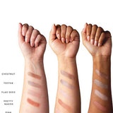Three forearms with various skin tones show swatches of the 100% Pure Pretty Naked Palette 9g—five vegan shades (Chestnut, Toffee, Flax Seed, Pretty Naked, Pink Champagne) arranged in rows from wrist to forearm.
