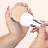 A person holds a compact of 100% Pure Rice Powder Foundation 9g in one hand and a large white makeup brush in the other, preparing to apply the talc-free pressed powder. Their manicured nails are painted with nude polish.