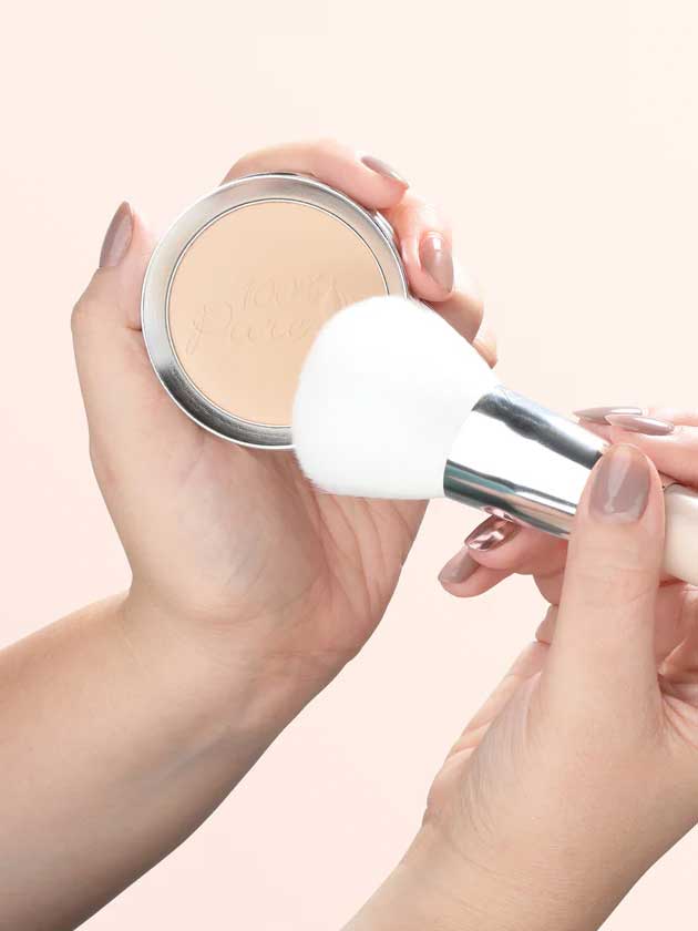 A person holds a compact of 100% Pure Rice Powder Foundation 9g in one hand and a large white makeup brush in the other, preparing to apply the talc-free pressed powder. Their manicured nails are painted with nude polish.