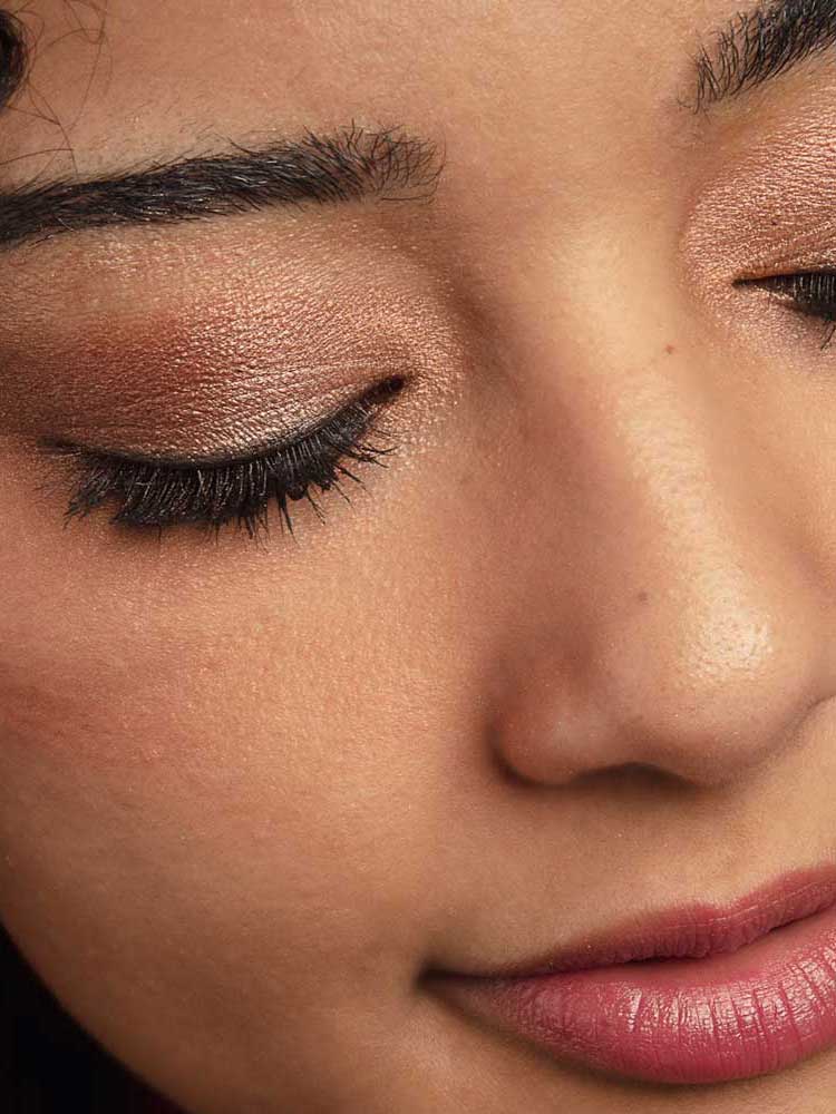 A close-up of a woman’s face featuring closed eyes adorned with shimmery brown from the 100% Pure Rose Gold Palette 9g, long dark lashes, smooth skin, and natural pink lips.