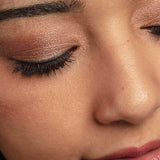 A close-up of a woman’s face featuring closed eyes adorned with shimmery brown from the 100% Pure Rose Gold Palette 9g, long dark lashes, smooth skin, and natural pink lips.