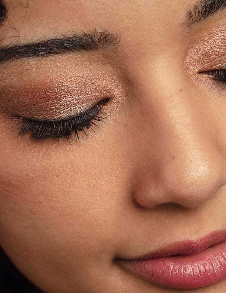 A close-up of a person’s face with smooth skin, closed eyes adorned in shimmery brown from the 100% Pure 100% Fruit Pigmented® Rose Gold Palette (9g), long lashes, and natural pink lips.