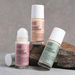Three Noosa Basics roll-on deodorants arranged around a rock: peach-labelled Coconut and Vanilla, green-labelled Lemon Myrtle, and pink-labelled Rose and Frankincense with its roller exposed and cap beside it.