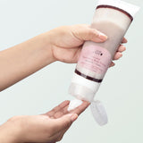A person holds a Coconut Nourishing Body Cream tube upside down above their other hand. Its flip-top lid hangs open as white cream is squeezed onto the waiting fingertips, against a pale mint background.