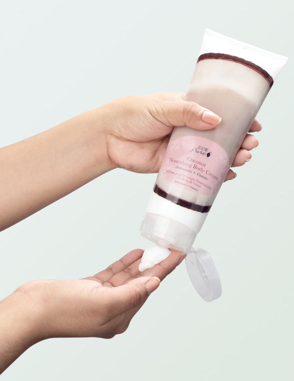 A person holds a Coconut Nourishing Body Cream tube upside down above their other hand. Its flip-top lid hangs open as white cream is squeezed onto the waiting fingertips, against a pale mint background.