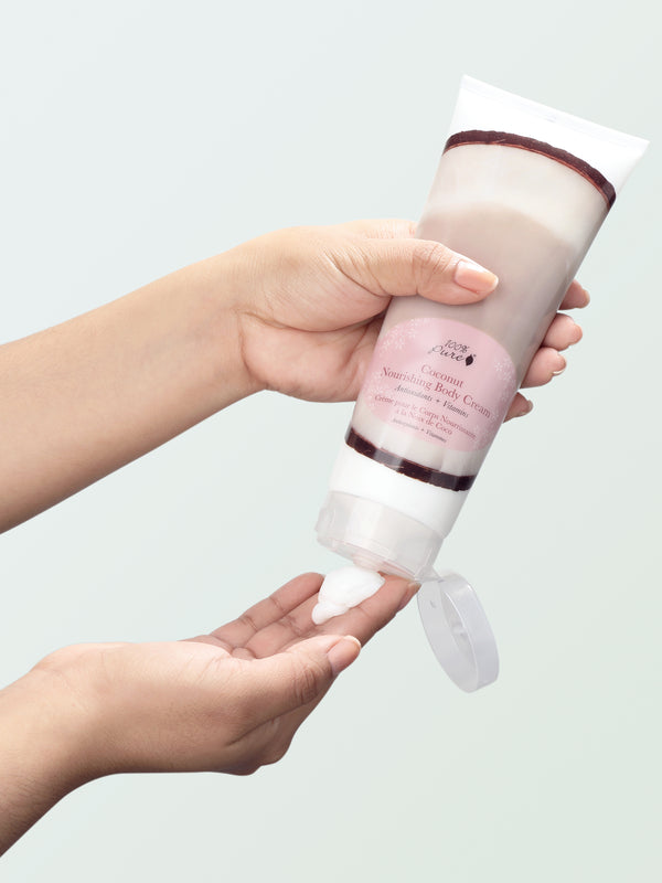 A person holds a Coconut Nourishing Body Cream tube upside down above their other hand. Its flip-top lid hangs open as white cream is squeezed onto the waiting fingertips, against a pale mint background.
