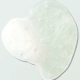 Close-up of a clear Coconut Shower Gel swatch on a pale mint surface. Its irregular outline surrounds fine bubbles and a foamy white patch concentrated on the left.
