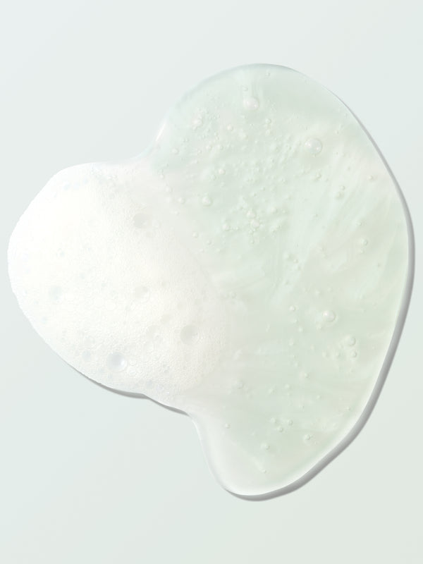 Close-up of a clear Coconut Shower Gel swatch on a pale mint surface. Its irregular outline surrounds fine bubbles and a foamy white patch concentrated on the left.