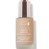 A closed frosted glass bottle of peach-beige 100% Pure 2nd Skin Serum Foundation has white lettering and a tall reflective rose-gold dropper cap. It casts a soft shadow to the right.