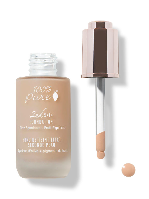 An open frosted bottle of peach-toned 100% Pure 2nd Skin Serum Foundation stands beside its rose-gold dropper. A coating of foundation clings to the pipette tip above a small drop on the white surface.