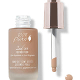 An open frosted bottle of tan 100% Pure 2nd Skin Serum Foundation stands beside its rose-gold dropper. Tan liquid coats the pipette tip, with a small round drop beneath it.