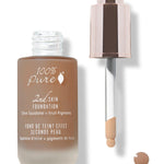 An open frosted bottle of brown 100% Pure 2nd Skin Serum Foundation stands beside its rose-gold dropper. The pipette tip is coated with foundation, with a small brown drop below it.