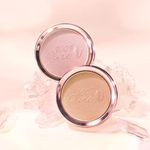 Two round 100% Pure luminiser pans, one shimmery pale pink and one warm gold, stand among translucent pink crystals against a softly lit peach background.