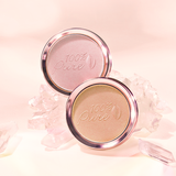 Two round 100% Pure luminiser pans, one shimmery pale pink and one warm gold, stand among translucent pink crystals against a softly lit peach background.
