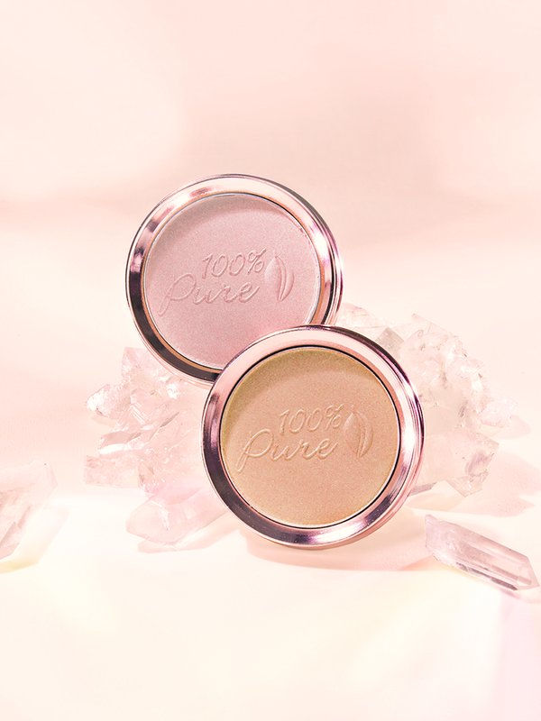 Two round 100% Pure luminiser pans, one shimmery pale pink and one warm gold, stand among translucent pink crystals against a softly lit peach background.