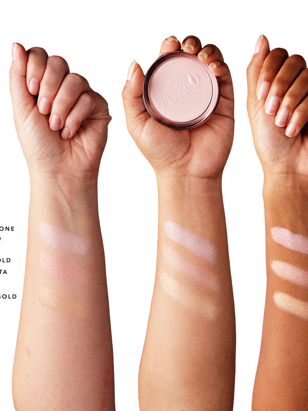 Three forearms with light, medium and dark skin each show three shimmering luminiser stripes: pearly pink nearest the wrist, rosy gold in the middle and warm gold below. The middle hand holds a pink powder compact; shade labels are cropped at the left edge.