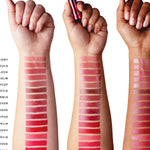 Three forearms with light, medium and dark skin each display 16 lipstick stripes, ranging from muted peach, rose and plum near the wrists to vivid pink, red and orange red towards the elbows. The middle hand holds a metallic pink lipstick tube. Shade names are cropped at the left edge.