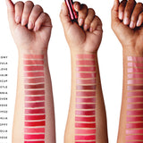 Three forearms with light, medium and dark skin each display 16 lipstick stripes, ranging from muted peach, rose and plum near the wrists to vivid pink, red and orange red towards the elbows. The middle hand holds a metallic pink lipstick tube. Shade names are cropped at the left edge.