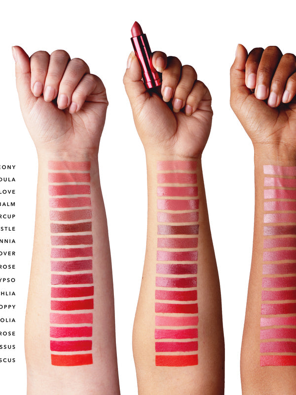Three forearms with light, medium and dark skin each display 16 lipstick stripes, ranging from muted peach, rose and plum near the wrists to vivid pink, red and orange red towards the elbows. The middle hand holds a metallic pink lipstick tube. Shade names are cropped at the left edge.