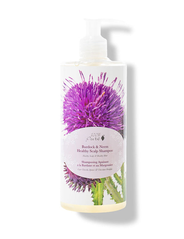A white pump bottle of 100% Pure Burdock and Neem Healthy Scalp Shampoo has a large purple thistle and green leaves behind a pale pink oval label. Pale yellow liquid is visible at the base.