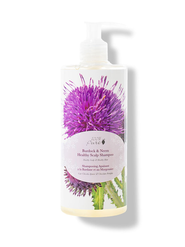A white pump bottle of 100% Pure Burdock and Neem Healthy Scalp Shampoo has a large purple thistle and green leaves behind a pale pink oval label. Pale yellow liquid is visible at the base.