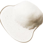 A translucent shampoo swatch forms a rounded irregular pool on white. Dense fine foam gathers at the upper right, with smaller bubbles scattered through the clear liquid.