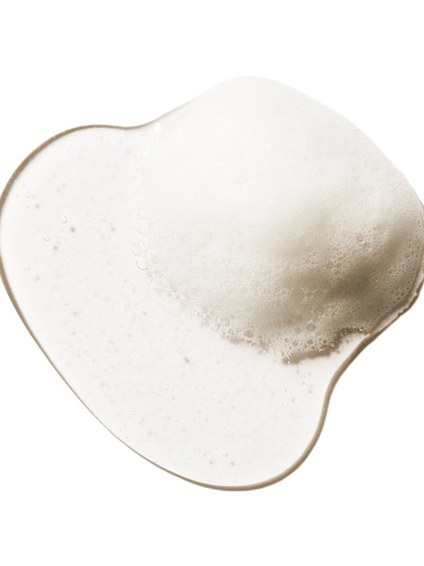 A translucent shampoo swatch forms a rounded irregular pool on white. Dense fine foam gathers at the upper right, with smaller bubbles scattered through the clear liquid.
