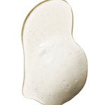 A pale, cloudy swatch of 100% Pure Yuzu & Pomelo Glossing Shampoo, forming a curved, bean-shaped puddle with fine foam bubbles and a glossy edge.