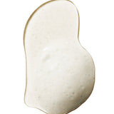 A pale, cloudy swatch of 100% Pure Yuzu & Pomelo Glossing Shampoo, forming a curved, bean-shaped puddle with fine foam bubbles and a glossy edge.