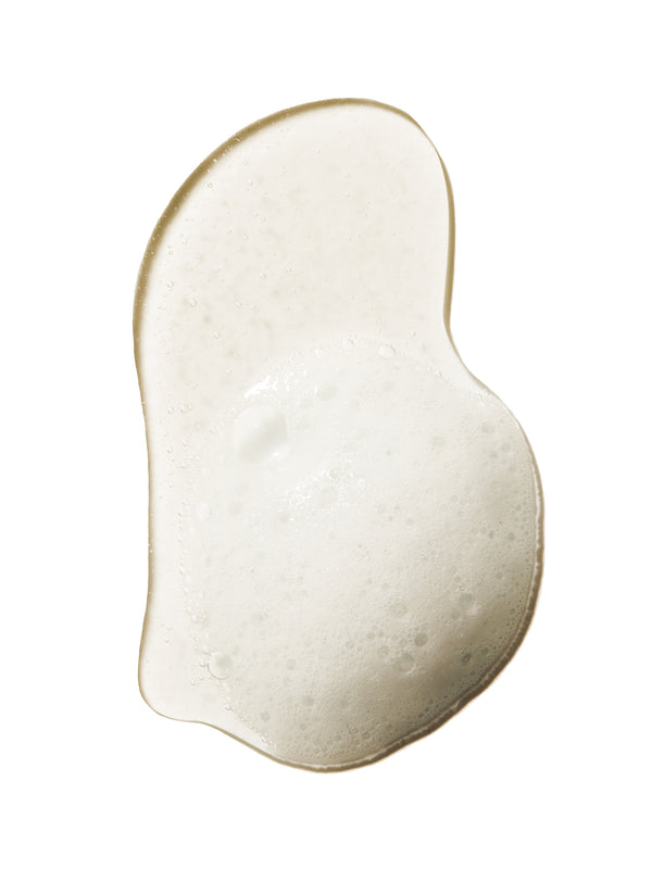 A pale, cloudy swatch of 100% Pure Yuzu & Pomelo Glossing Shampoo, forming a curved, bean-shaped puddle with fine foam bubbles and a glossy edge.