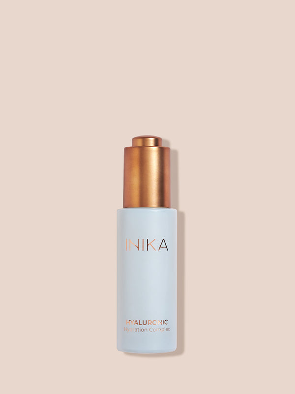 Pale blue INIKA Hyaluronic Hydration Complex bottle standing upright with its gold pump exposed on a beige background, casting a narrow shadow to the right.