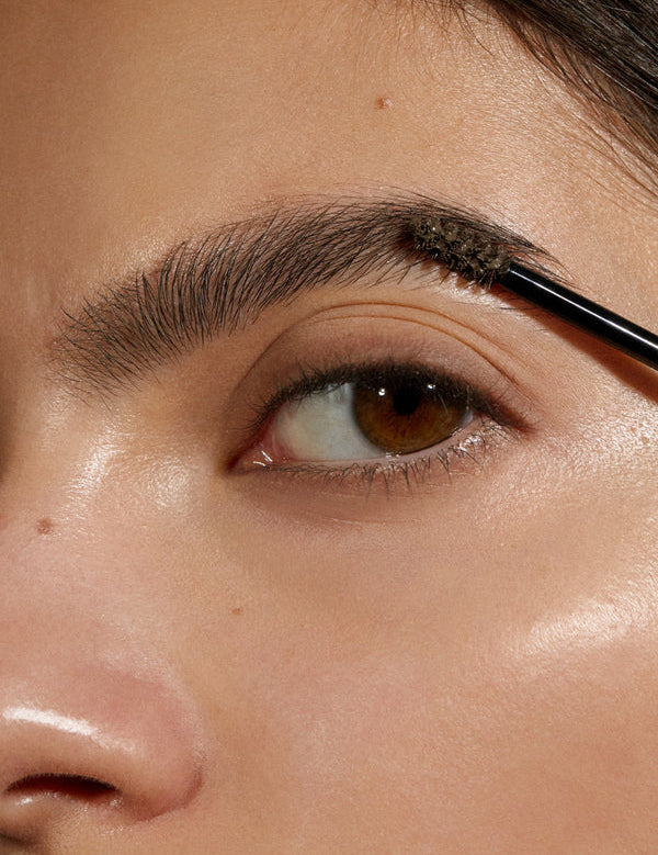 Close-up of a woman's brown eye and eyebrow as a small brush combs Madara Grow & Fix tinted brow gel in Ash Brown through the brow hairs.