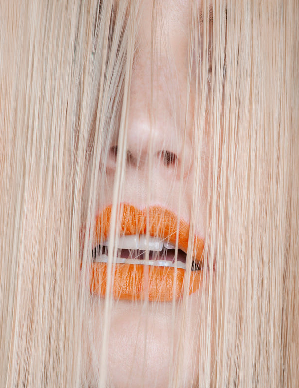 Close-up of a woman's face behind a curtain of fine, straight blonde hair, with bright orange lips visible through the strands.