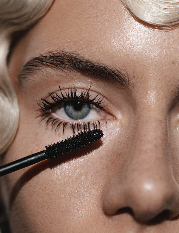 Close-up of a blue eye with long, separated black lashes and a black Madara Deep Matter mascara brush held just below the lower lashes.
