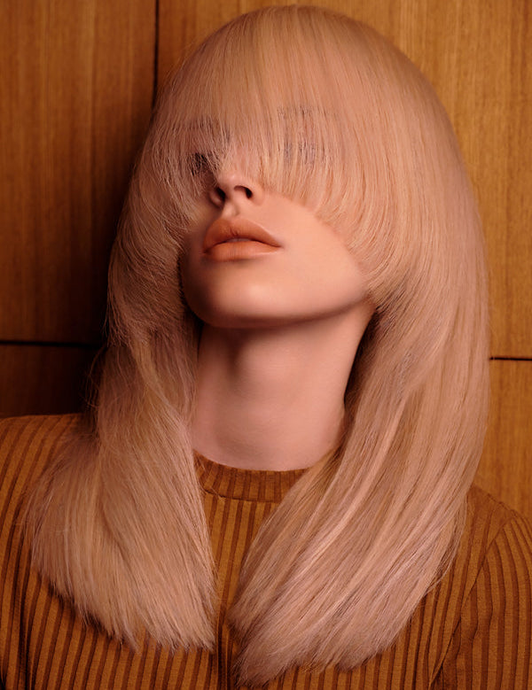 Woman with long, smooth peach-blonde hair curving over both eyes and around her shoulders, wearing a brown striped top against wood panelling.