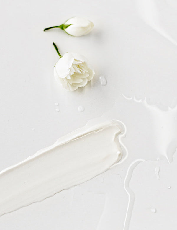 A long smear of white Madara Cleansing Milk on a white surface, with two small white rosebuds and scattered clear droplets above it.
