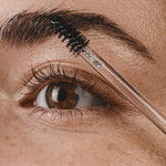 Close-up of a brown eye and full eyebrow as a black spiral brush on a clear wand combs Madara Grow Fix clear booster through the brow hairs.