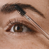 Close-up of a brown eye and full eyebrow as a black spiral brush on a clear wand combs Madara Grow Fix clear booster through the brow hairs.