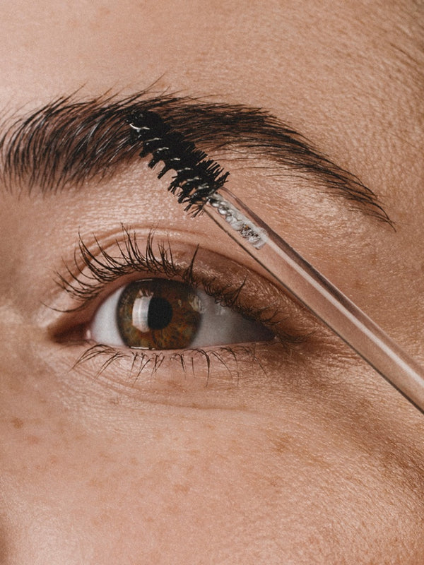 Close-up of a brown eye and full eyebrow as a black spiral brush on a clear wand combs Madara Grow Fix clear booster through the brow hairs.