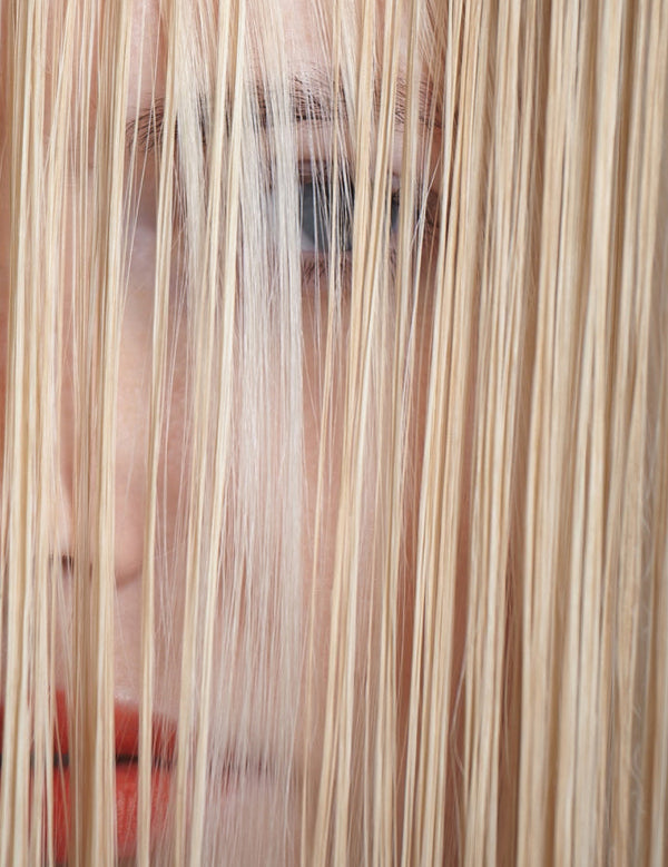 Close-up of a woman's face partly hidden behind long, straight blonde strands, with one eye and her red lips visible through the hair.