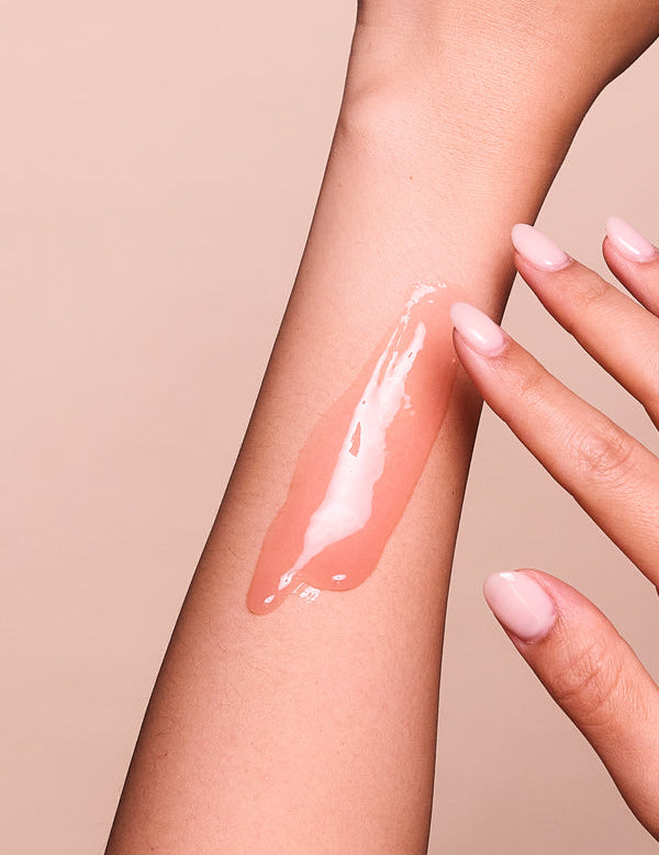 Fingers spreading a long, glossy streak of translucent pink Jojoba and Enzyme Radiance Mask along the inner forearm against a peach-pink background.