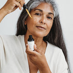 Woman with long grey hair applying Mukti Vital A Vitamin Booster to her face with the dropper