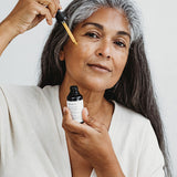 Woman with long grey hair applying Mukti Vital A Vitamin Booster to her face with the dropper